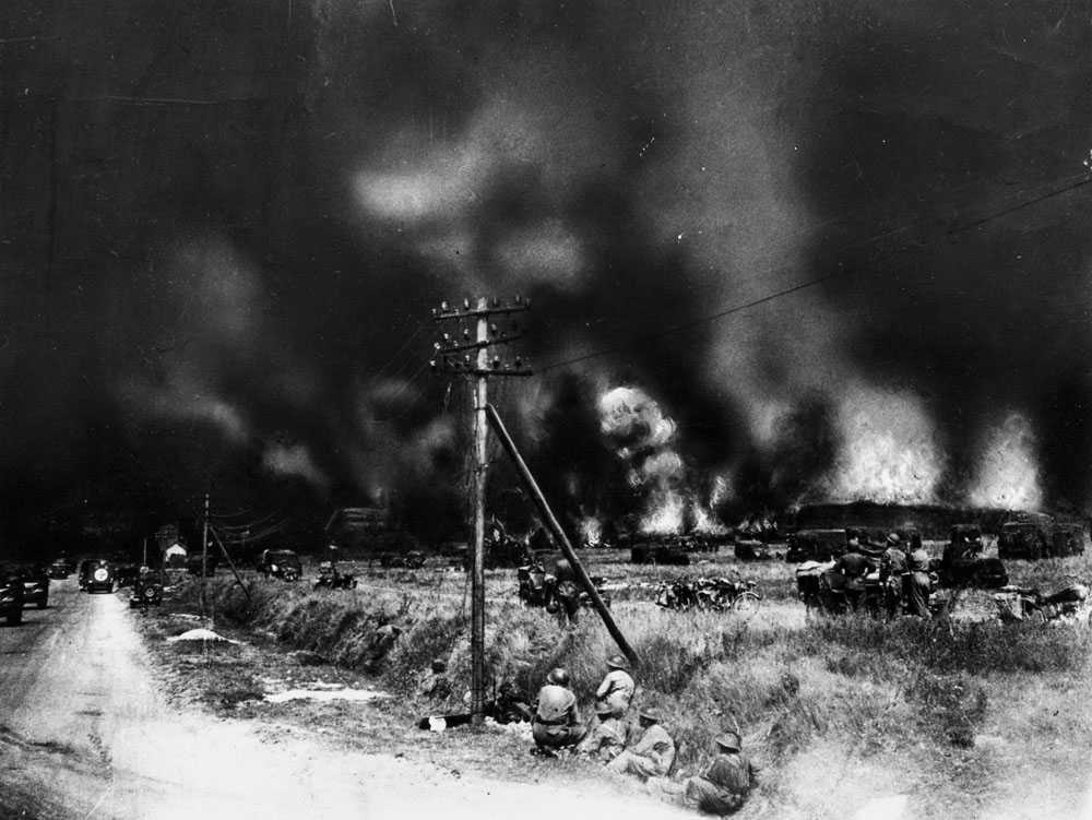 Photographie en noir et blanc – Une route et un champ ouvert. De la fumée et des explosions sont visibles au loin.